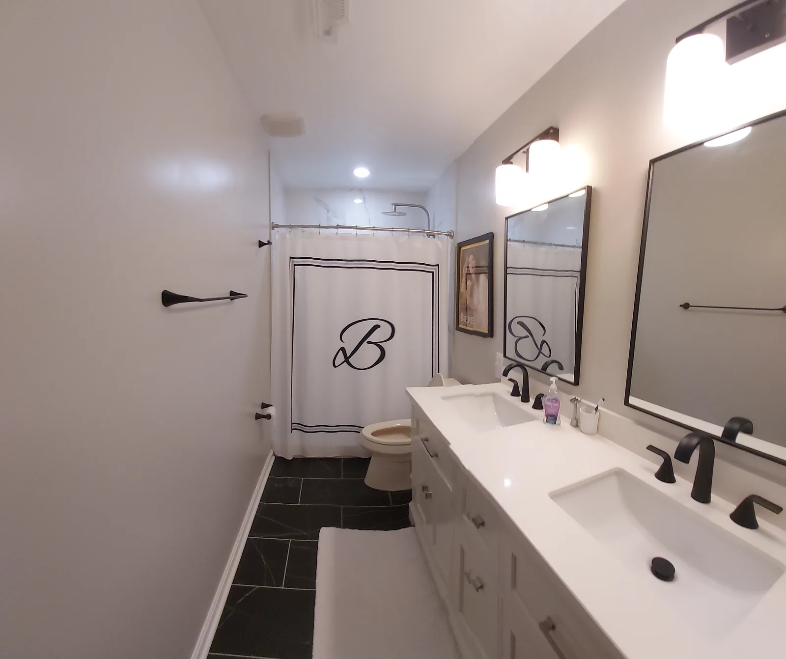 Long bathroom with a white double vanity, two black framed mirrors and light fixtures, black faucets, a dark stone patterned floor, and a shower curtain at the far end.