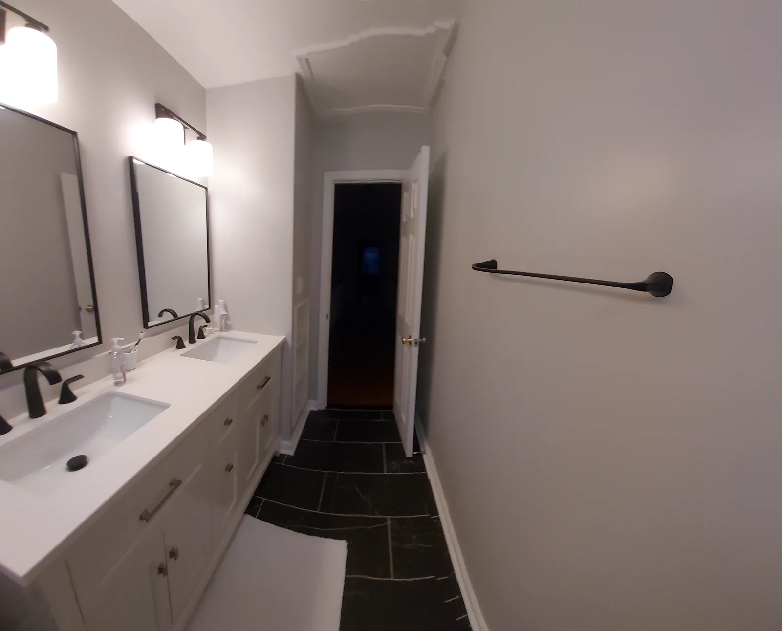 White double vanity with cabinet doors and drawers, black faucets, separate framed mirrors, a dark stone patterned floor, and a towel bar on the opposite wall.