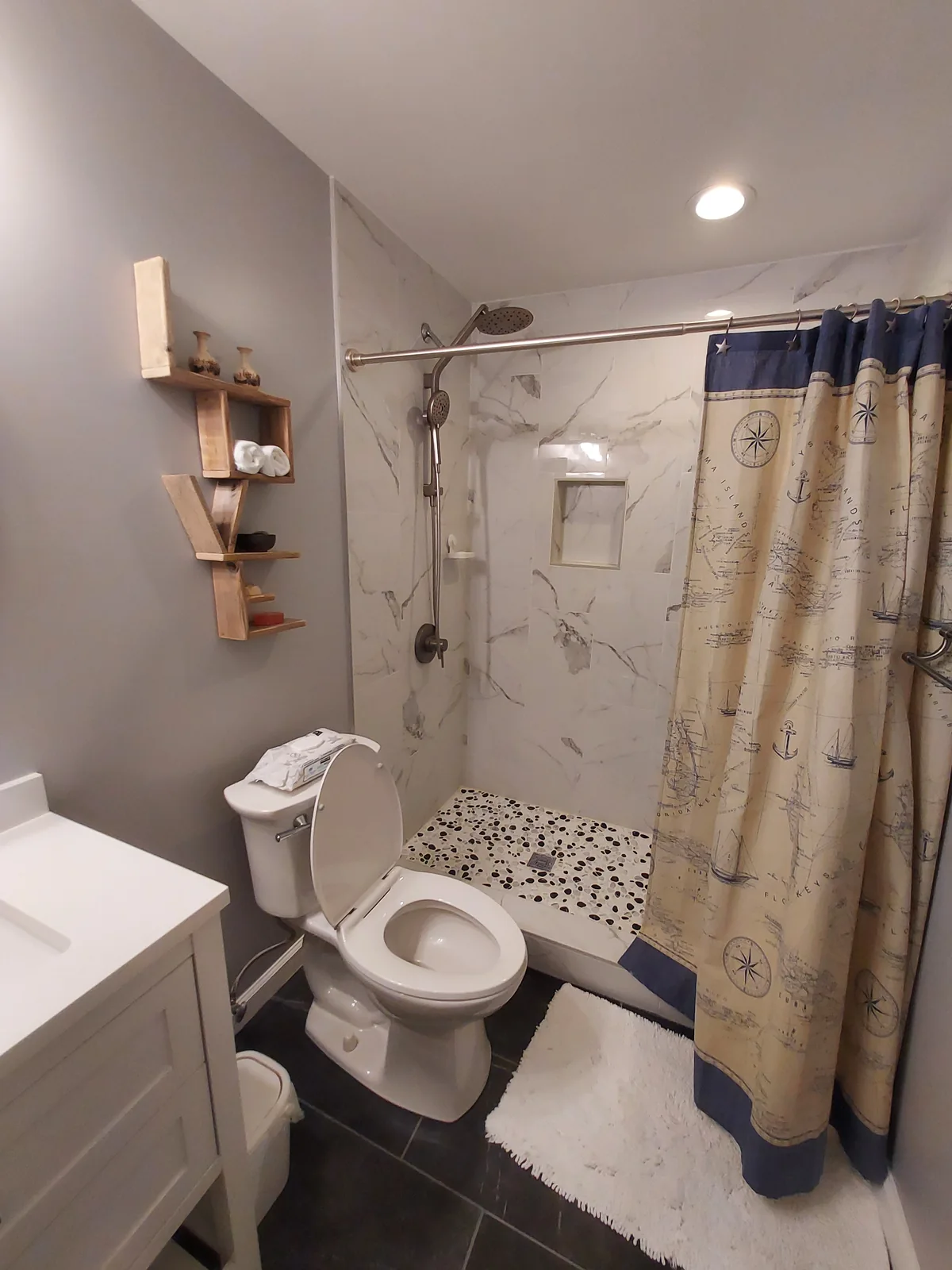 Compact bathroom with a shower featuring white stone patterned walls, overhead and handheld showerheads, a recessed niche, a pebble patterned floor, and a curtain beside dark bathroom flooring.
