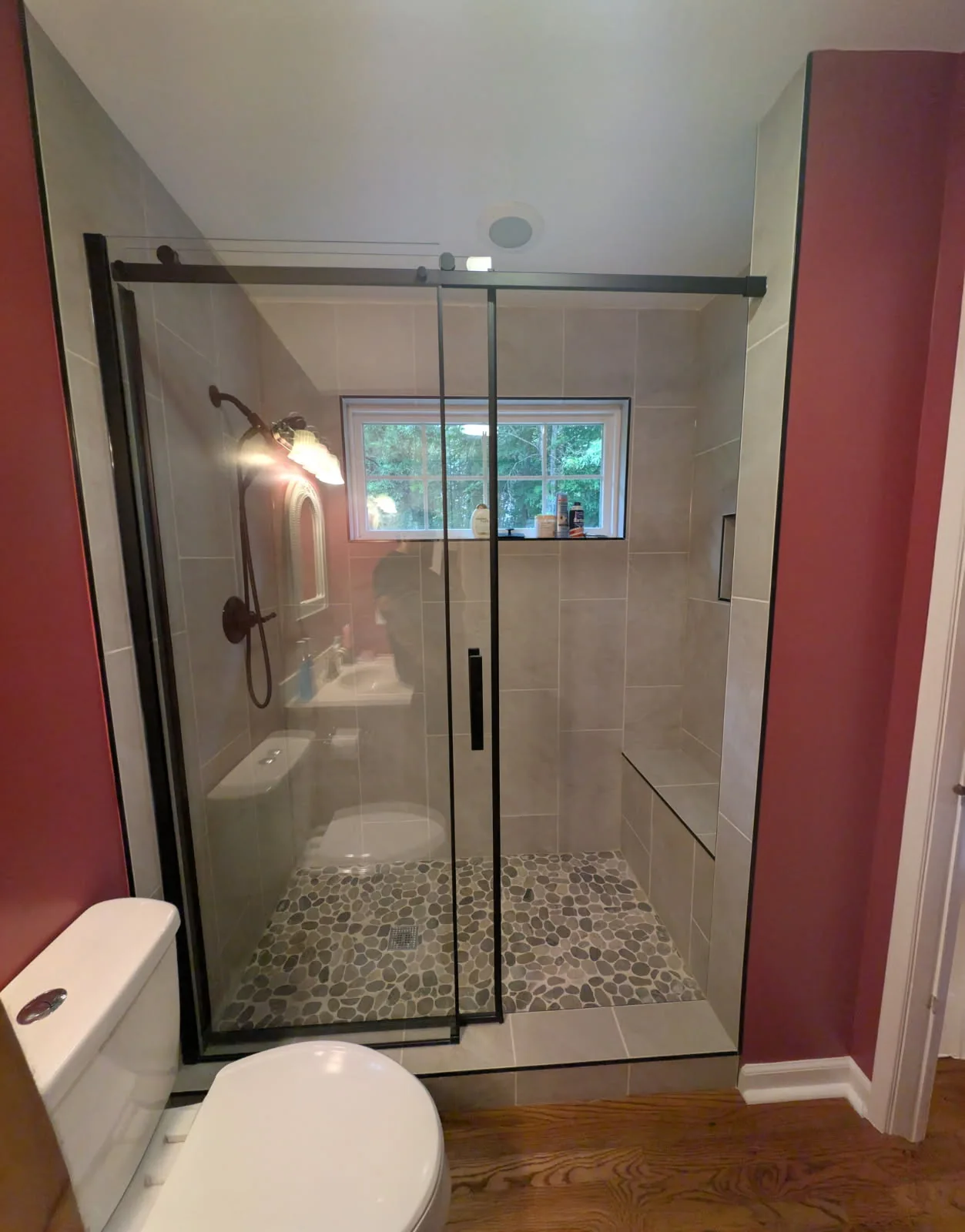 Shower behind sliding glass doors with black hardware, pale wall finishes, a pebble patterned floor, a built in bench, a recessed niche, and a window.